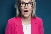 Blonde Frau mit schwarzer Brille und pinkfarbenem Blazer vor dunkelblauem Hintergrund.