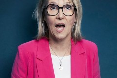 Blonde Frau mit schwarzer Brille und pinkfarbenem Blazer vor dunkelblauem Hintergrund.