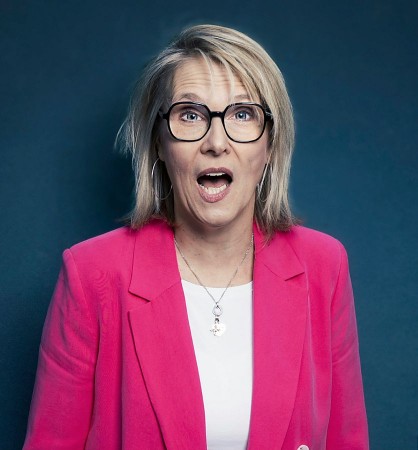 Blonde Frau mit schwarzer Brille und pinkfarbenem Blazer vor dunkelblauem Hintergrund.