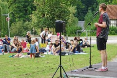 Poetry-Slammer auf einer kleinen Freiluftbühne vor einem auf Decken sitzenden Publikum auf einer Wiese.