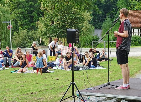 Poetry-Slammer auf einer kleinen Freiluftbühne vor einem auf Decken sitzenden Publikum auf einer Wiese.