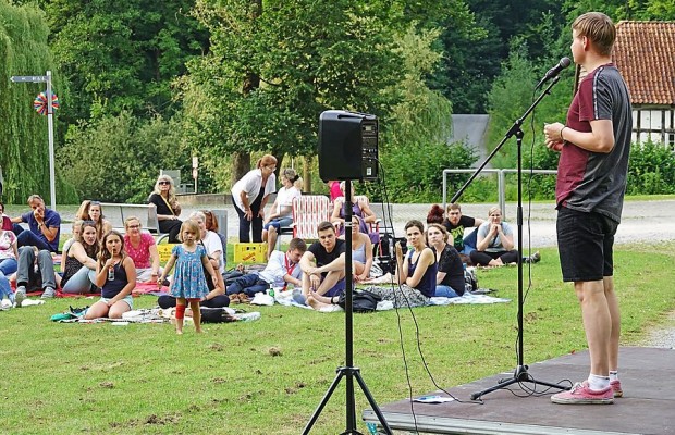 Poetry-Slammer auf einer kleinen Freiluftbühne vor einem auf Decken sitzenden Publikum auf einer Wiese.
