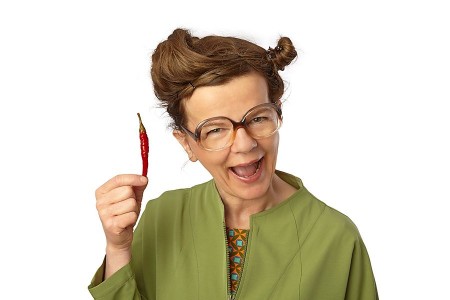 Frau in grüner Jacke hält eine rote Chilischote neben ihr Gesicht.