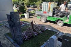 Mitarbeiter eines friedhofsgärtnerischen Betriebs mit einem Bewässerungsfahrzeug zwischen bepflanzten Gräbern auf einem Friedhof.