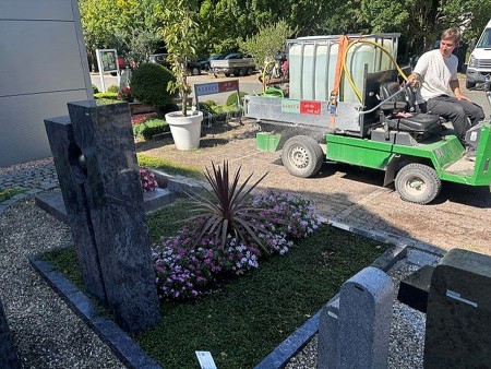 Mitarbeiter eines friedhofsgärtnerischen Betriebs mit einem Bewässerungsfahrzeug zwischen bepflanzten Gräbern auf einem Friedhof.