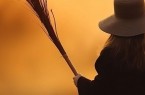 Frau mit breitkrempigem Hut hält einen Reisigbesen vor orangefarbenem Hintergrund.