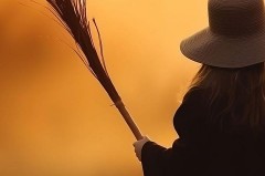 Frau mit breitkrempigem Hut hält einen Reisigbesen vor orangefarbenem Hintergrund.