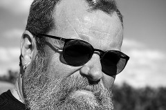 Schwarz-Weiß-Porträt eines bärtigen Mannes mit Sonnenbrille im Freien.