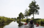 Zwei Personen fahren in einem Kanu auf einem ruhigen Fluss zwischen bewachsenen Ufern und Bäumen.