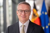 Porträt eines Mannes im Anzug vor unscharf dargestellter deutscher und europäischer Flagge.