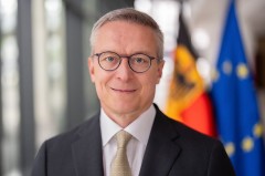Porträt eines Mannes im Anzug vor unscharf dargestellter deutscher und europäischer Flagge.