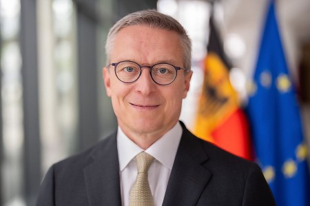 Porträt eines Mannes im Anzug vor unscharf dargestellter deutscher und europäischer Flagge.