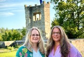 Zwei Frauen stehen auf einer Grünfläche vor einem steinernen Turm mit Europaflagge.