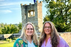 Zwei Frauen stehen auf einer Grünfläche vor einem steinernen Turm mit Europaflagge.
