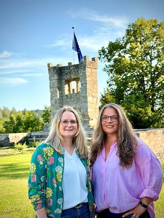 Zwei Frauen stehen auf einer Grünfläche vor einem steinernen Turm mit Europaflagge.