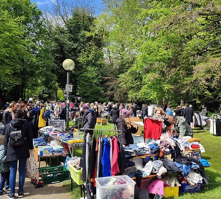 Gut besuchter Flohmarkt im Grünen mit Kleiderständern, Tischen, Spielzeug und zahlreichen Besucherinnen und Besuchern.