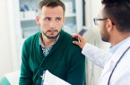 Arzt legt einem Mann im grünen Pullover während eines Gesprächs die Hand auf die Schulter.