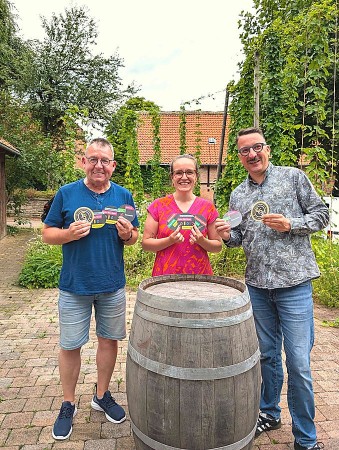 Drei Personen mit bedruckten Bierdeckeln der Bürgerbrauzunft Nieheim vor einem Holzfass.