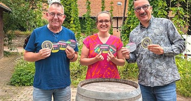 Drei Personen mit bedruckten Bierdeckeln der Bürgerbrauzunft Nieheim vor einem Holzfass.