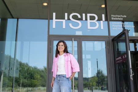Eine Frau steht vor dem Eingang der Hochschule Bielefeld unter dem großen HSBI-Schriftzug.