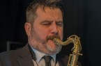 Ein bärtiger Saxophonist im Anzug spielt mit geschlossenen Augen auf einer Bühne.