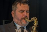 Ein bärtiger Saxophonist im Anzug spielt mit geschlossenen Augen auf einer Bühne.