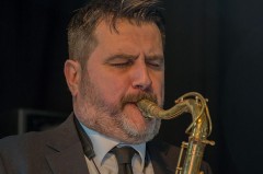 Ein bärtiger Saxophonist im Anzug spielt mit geschlossenen Augen auf einer Bühne.
