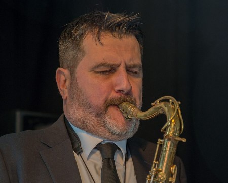 Ein bärtiger Saxophonist im Anzug spielt mit geschlossenen Augen auf einer Bühne.