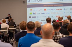 Publikum bei einer SICP-Veranstaltung vor einer Präsentation mit Logos von Mitgliedsunternehmen.