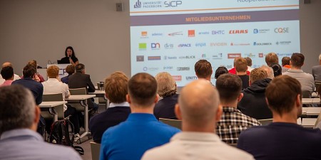 Publikum bei einer SICP-Veranstaltung vor einer Präsentation mit Logos von Mitgliedsunternehmen.