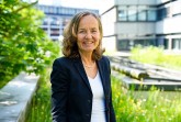 Porträt einer lächelnden Frau in dunklem Blazer vor einem modernen Gebäude und einer begrünten Außenanlage.