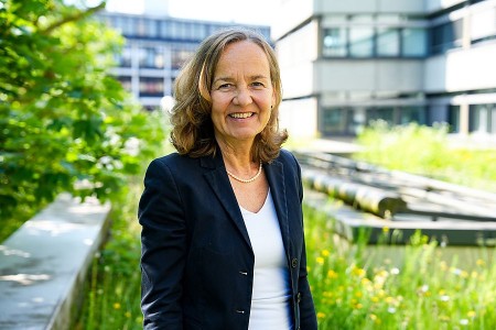 Porträt einer lächelnden Frau in dunklem Blazer vor einem modernen Gebäude und einer begrünten Außenanlage.