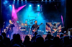 Vierköpfige Band ReCartney spielt mit Gitarren und Schlagzeug auf einer blau und pink beleuchteten Bühne vor Publikum.