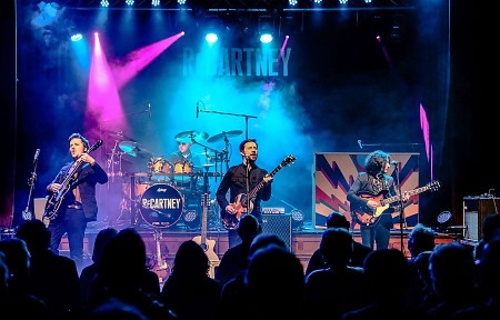 Vierköpfige Band ReCartney spielt mit Gitarren und Schlagzeug auf einer blau und pink beleuchteten Bühne vor Publikum.