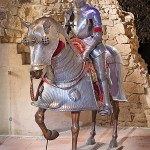 Ritterfigur in vollständiger Metallrüstung sitzt auf einem gepanzerten Pferdemodell vor einer historischen Steinwand.