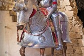 Ritterfigur in vollständiger Metallrüstung sitzt auf einem gepanzerten Pferdemodell vor einer historischen Steinwand.
