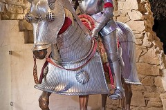 Ritterfigur in vollständiger Metallrüstung sitzt auf einem gepanzerten Pferdemodell vor einer historischen Steinwand.