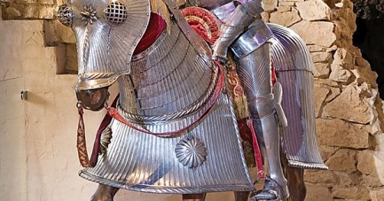 Ritterfigur in vollständiger Metallrüstung sitzt auf einem gepanzerten Pferdemodell vor einer historischen Steinwand.