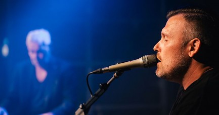 Sebastian Hegener singt bei einem Auftritt in ein Mikrofon und spielt Gitarre.