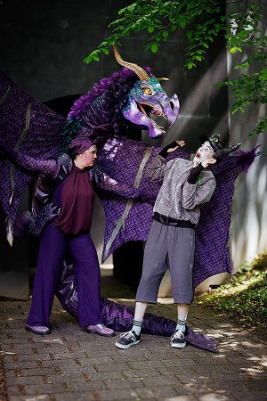Ein großer violetter Drache beugt sich über den erschrockenen Esel, während eine Darstellerin in violettem Kostüm danebensteht.