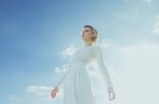 Stina Holmquist in weißem Outfit vor blauem Himmel, vertikales Porträt aus niedriger Perspektive.