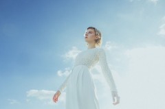 Stina Holmquist in weißem Outfit vor blauem Himmel, vertikales Porträt aus niedriger Perspektive.