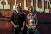 Zwei Männer mit Cowboyhüten und Sonnenbrillen stehen vor einer roten Wand mit großem Saloon-Schriftzug.