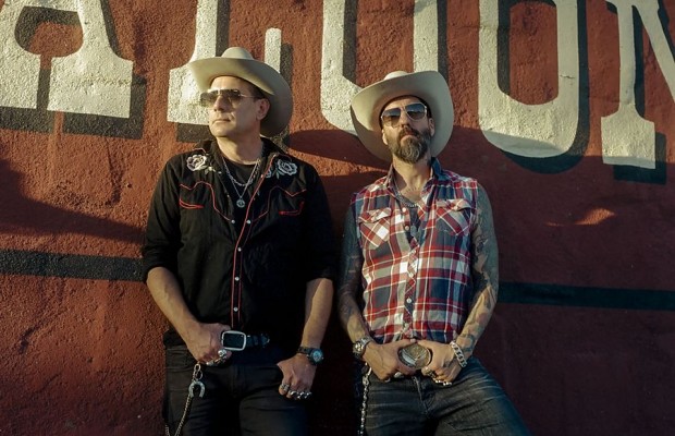 Zwei Männer mit Cowboyhüten und Sonnenbrillen stehen vor einer roten Wand mit großem Saloon-Schriftzug.
