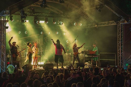 Musikerinnen und Musiker auf der beleuchteten Bühne des Turnpike Reggae Festivals vor einem dicht stehenden Publikum.