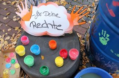 Bunte Fingerfarben, orange Handformen und ein Schild mit der Aufschrift „für Deine Rechte“ neben bemalten Tonnen.