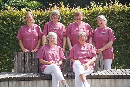 Sechs Mitarbeiterinnen des Wohnzimmer-Projekts im Klinikum Gütersloh in rosa Pflegekleidung vor einer Hecke.