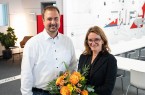 Ein Mann im weißen Hemd und eine Frau im schwarzen Blazer stehen mit einem Blumenstrauß in einem modernen Besprechungsraum.