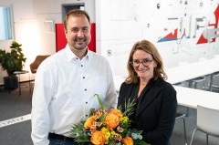 Ein Mann im weißen Hemd und eine Frau im schwarzen Blazer stehen mit einem Blumenstrauß in einem modernen Besprechungsraum.
