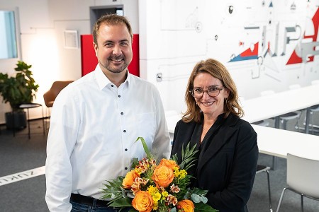 Ein Mann im weißen Hemd und eine Frau im schwarzen Blazer stehen mit einem Blumenstrauß in einem modernen Besprechungsraum.